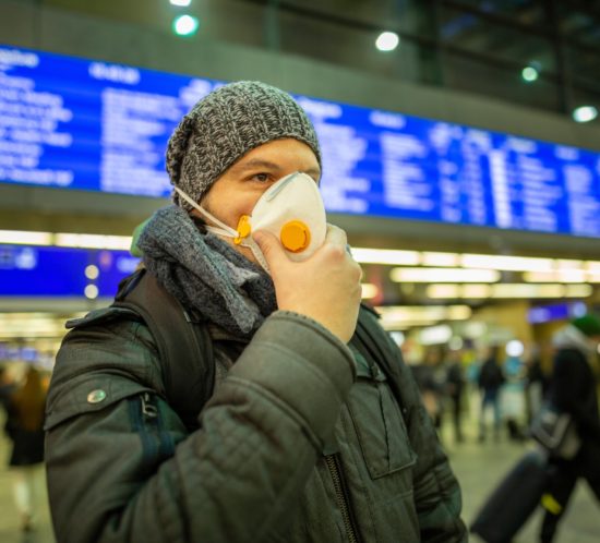 Man wearing a respirator mask device for health protection at an airport or railway train station in a crowd of people while travelling. Epidemic corona virus infection, flu sickness and travel illness concept.